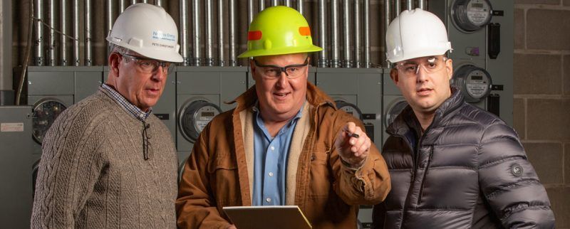 Three construction workers in hard hats discuss a project. One points while holding a clipboard.