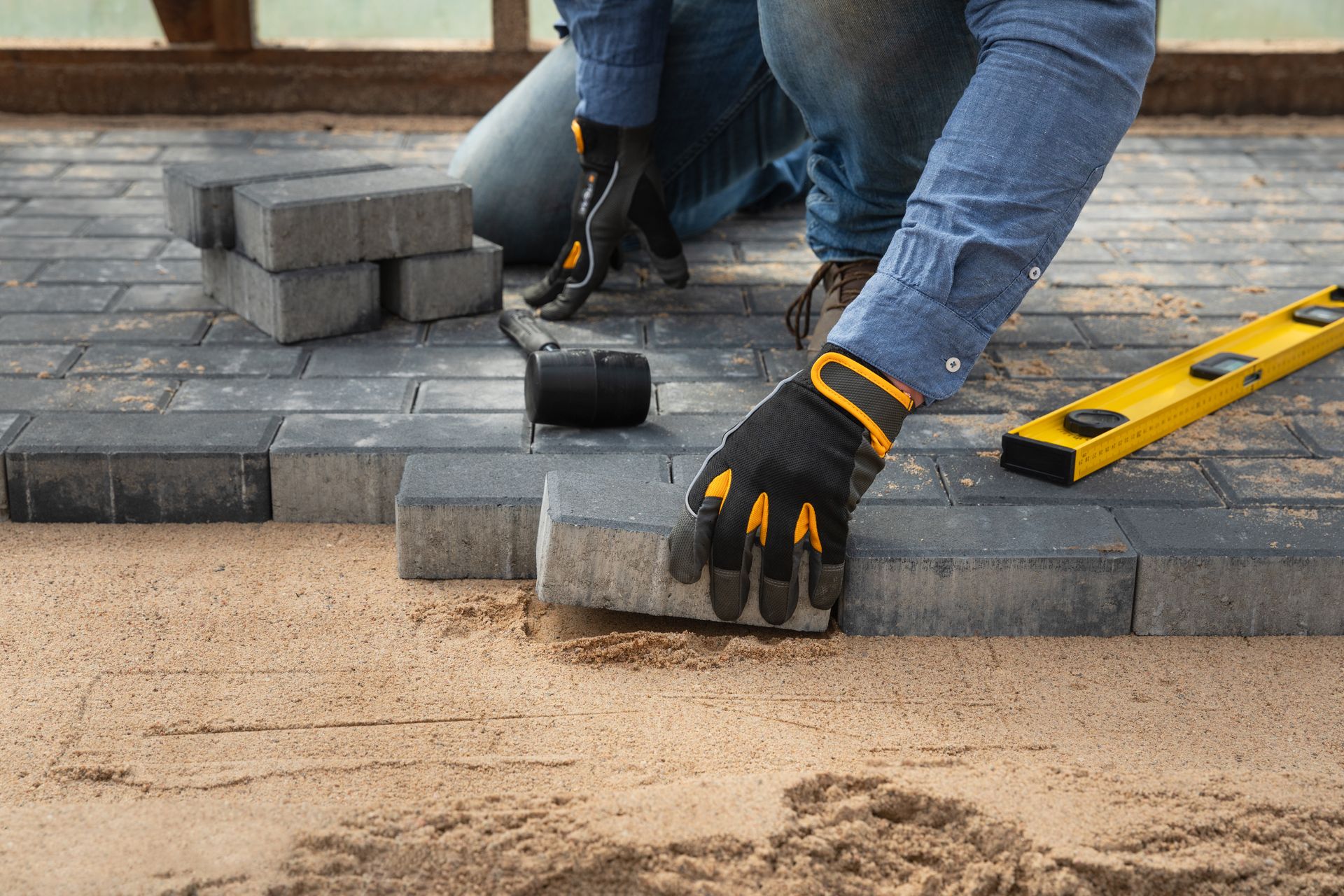 Worker using a rubber hammer and gloves to lay cement pavement on a sandy walkway.