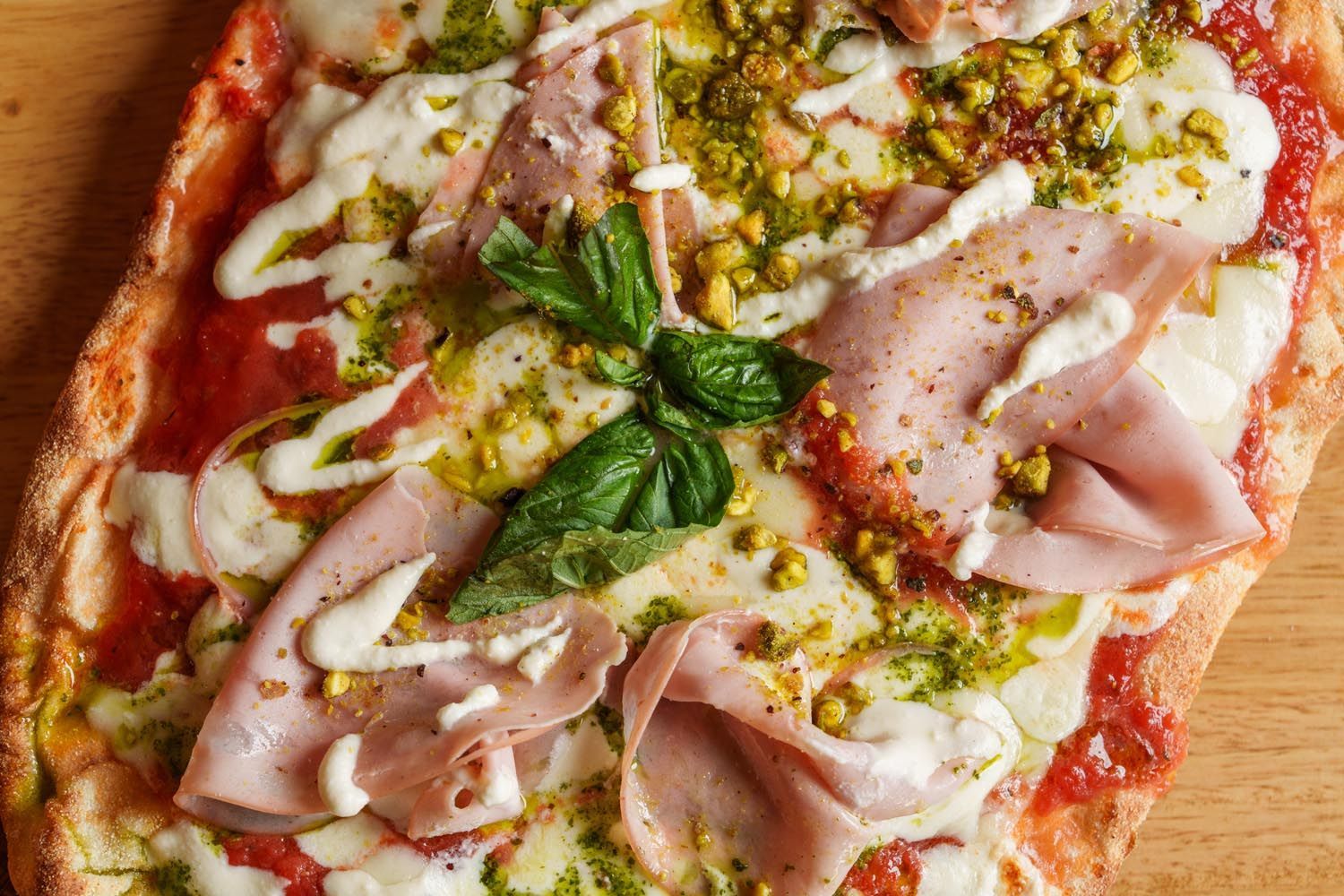 A pizza with ham , cheese , basil and pistachios on a wooden table.