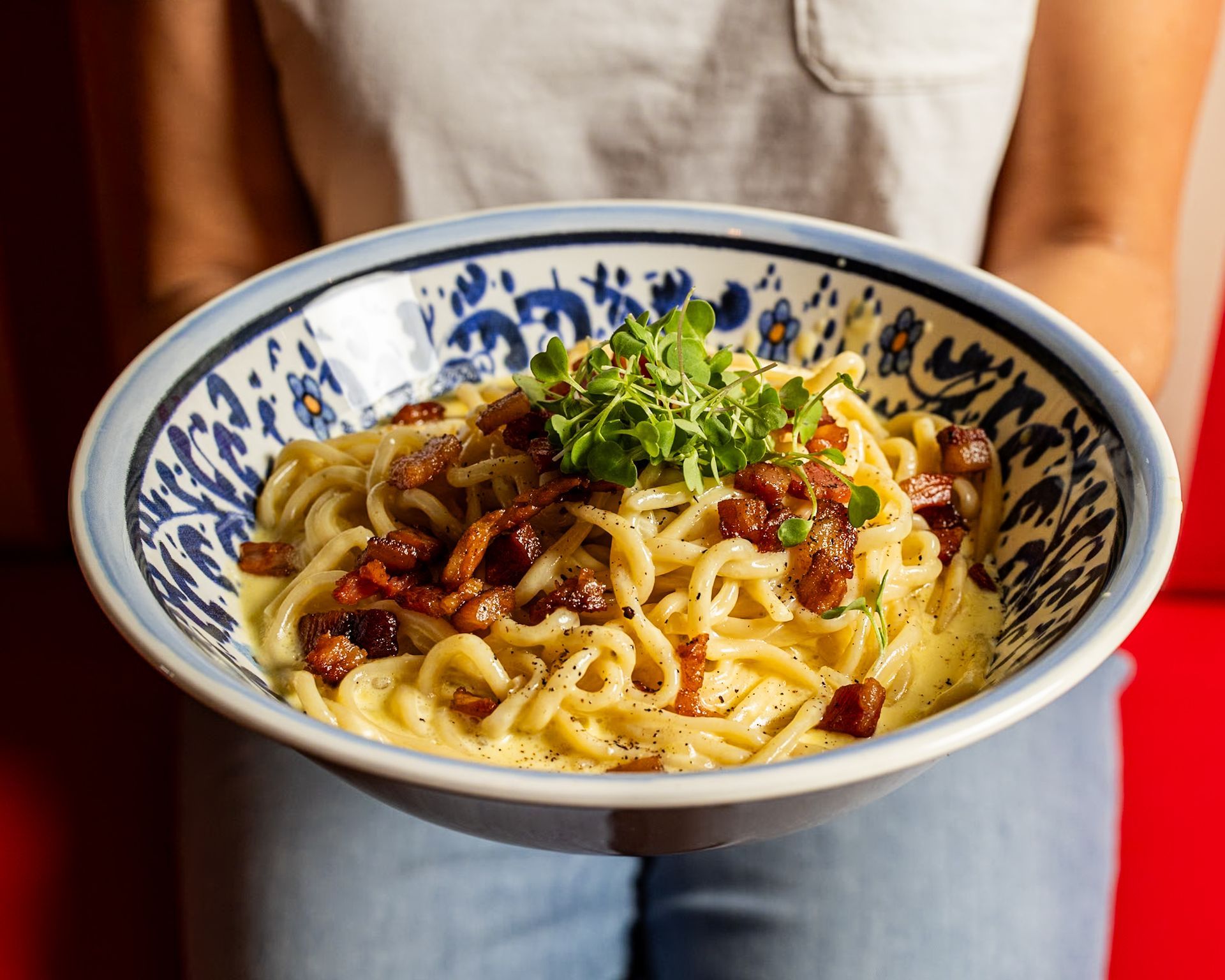 A woman is holding a bowl of pasta with bacon and parsley.