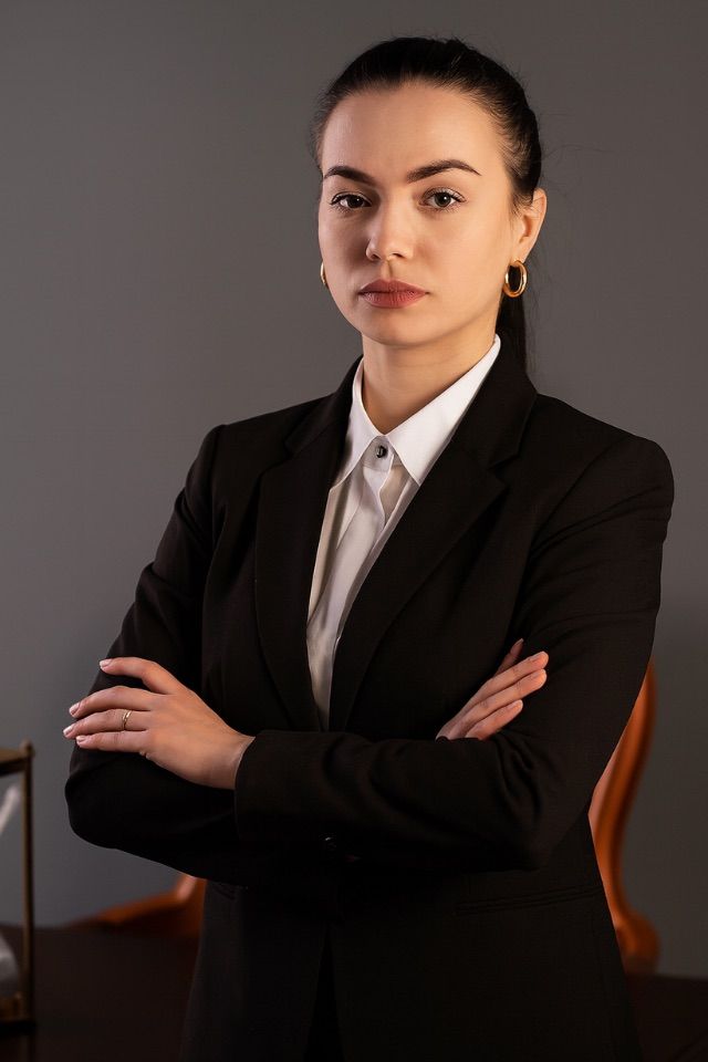 Mujer con traje negro, brazos cruzados y expresión seria. De pie en una oficina.