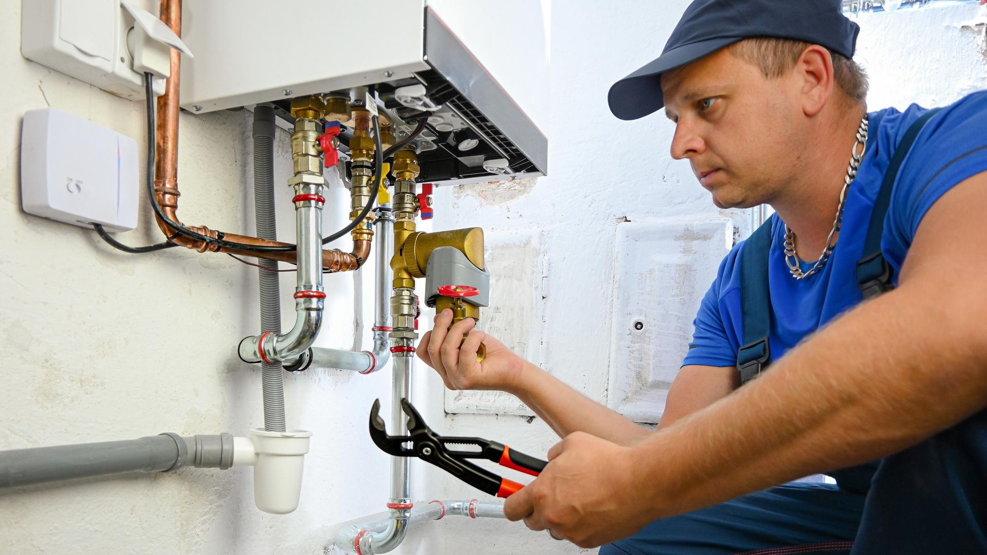 Plumber in blue overalls and cap working on a boiler, using pliers, indoor setting. Plumber in blue overalls and cap working on a boiler, using pliers, indoor setting.