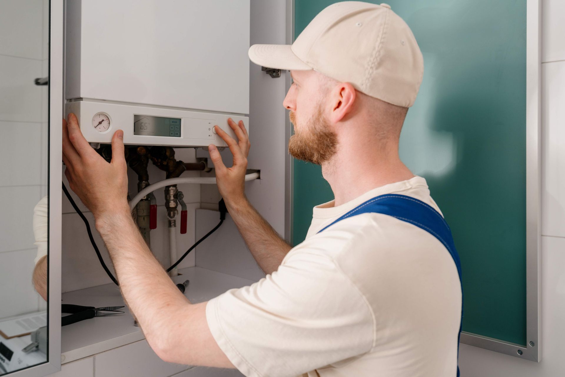 Man in work clothes fixing a white boiler mounted on a wall.