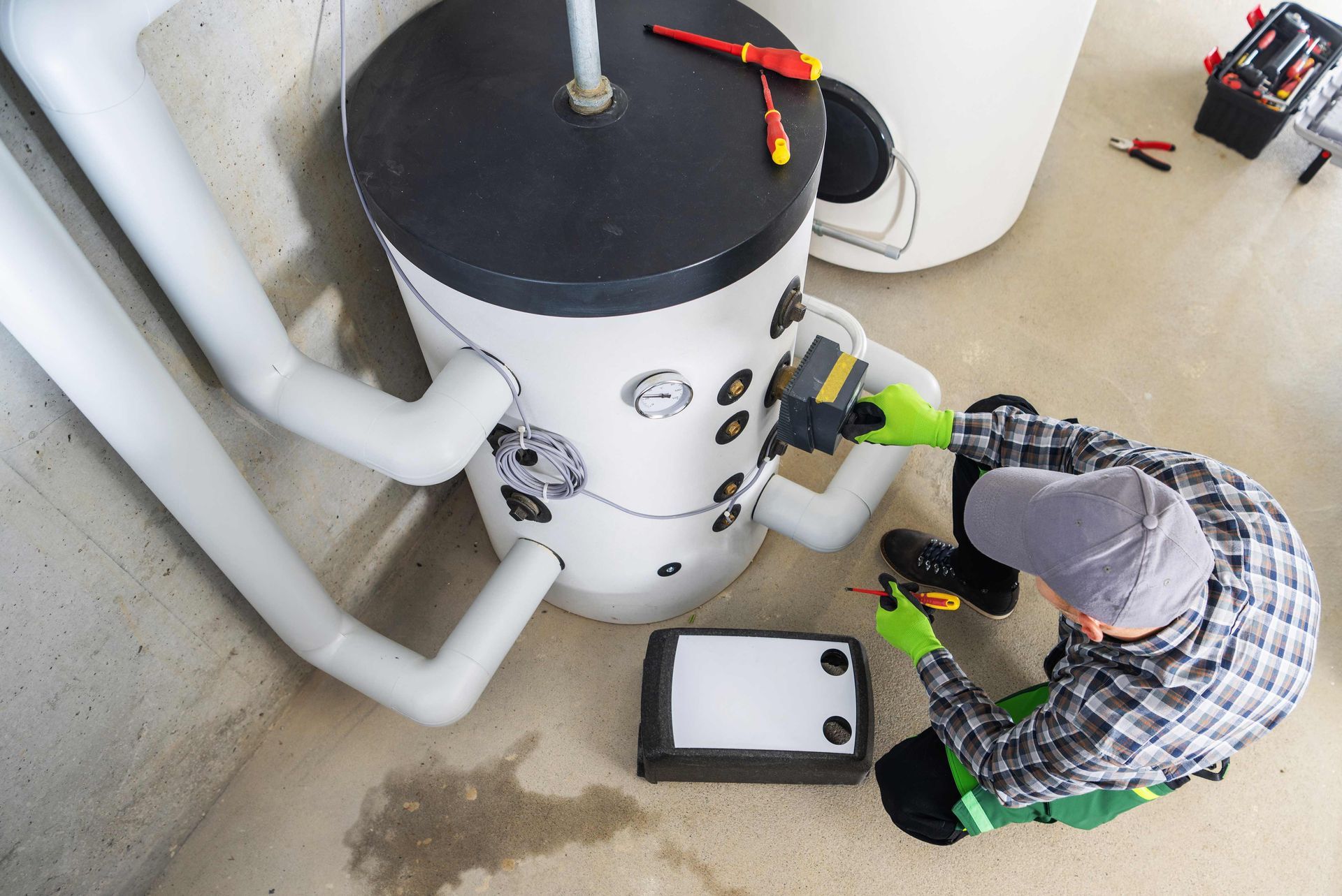 Plumber in green gloves repairs a water heater in a basement, using a screwdriver.