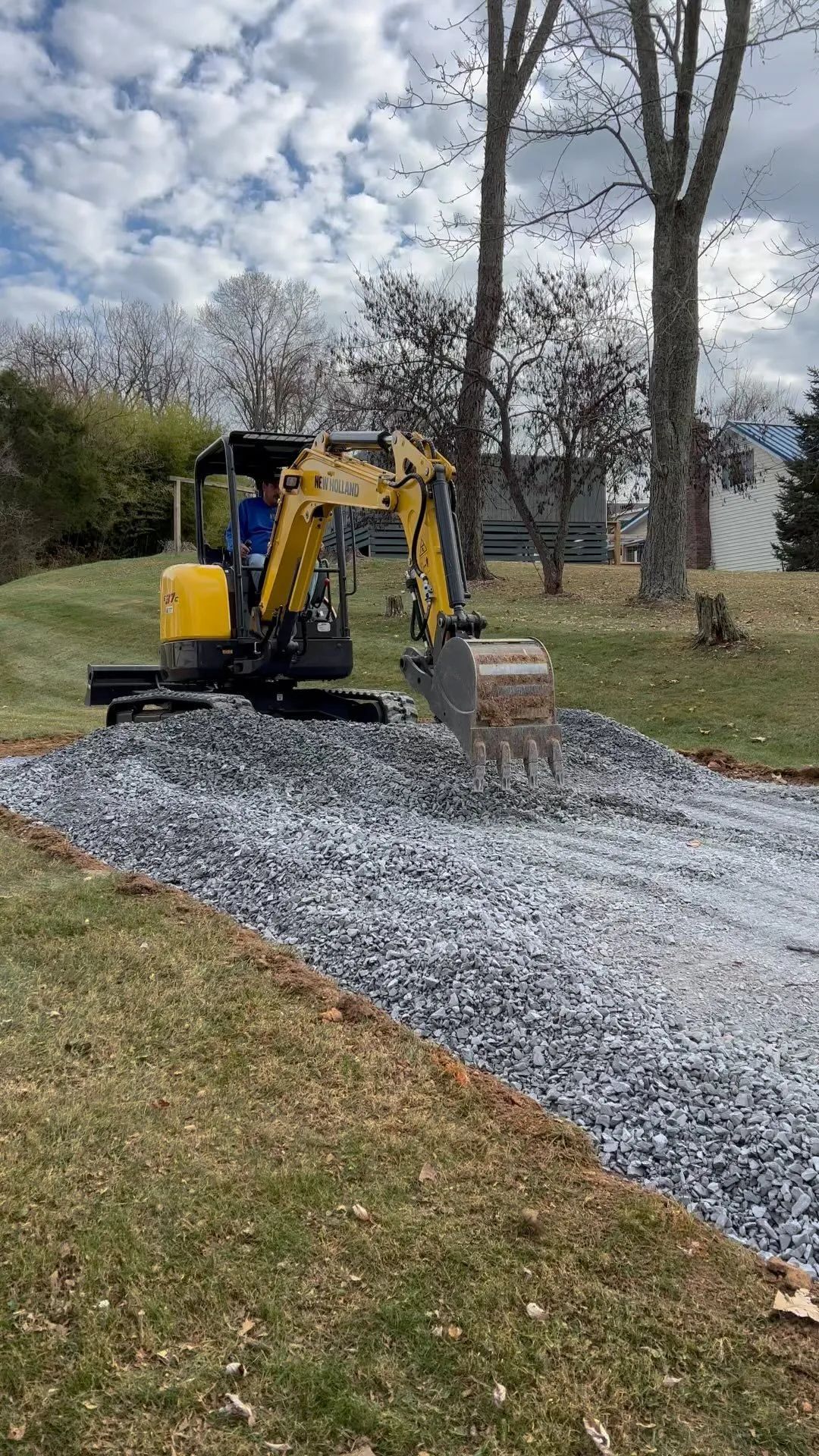 A yellow excavator on a gravel driveway. A person is operating the machine. Trees and a house in the background.