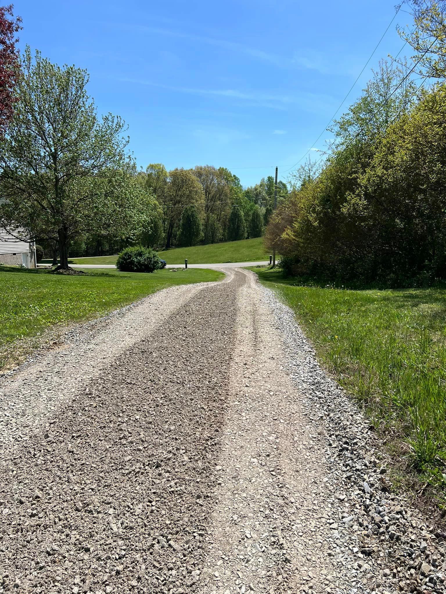 Gravel driveway through a grassy area, trees on either side, under a blue sky.