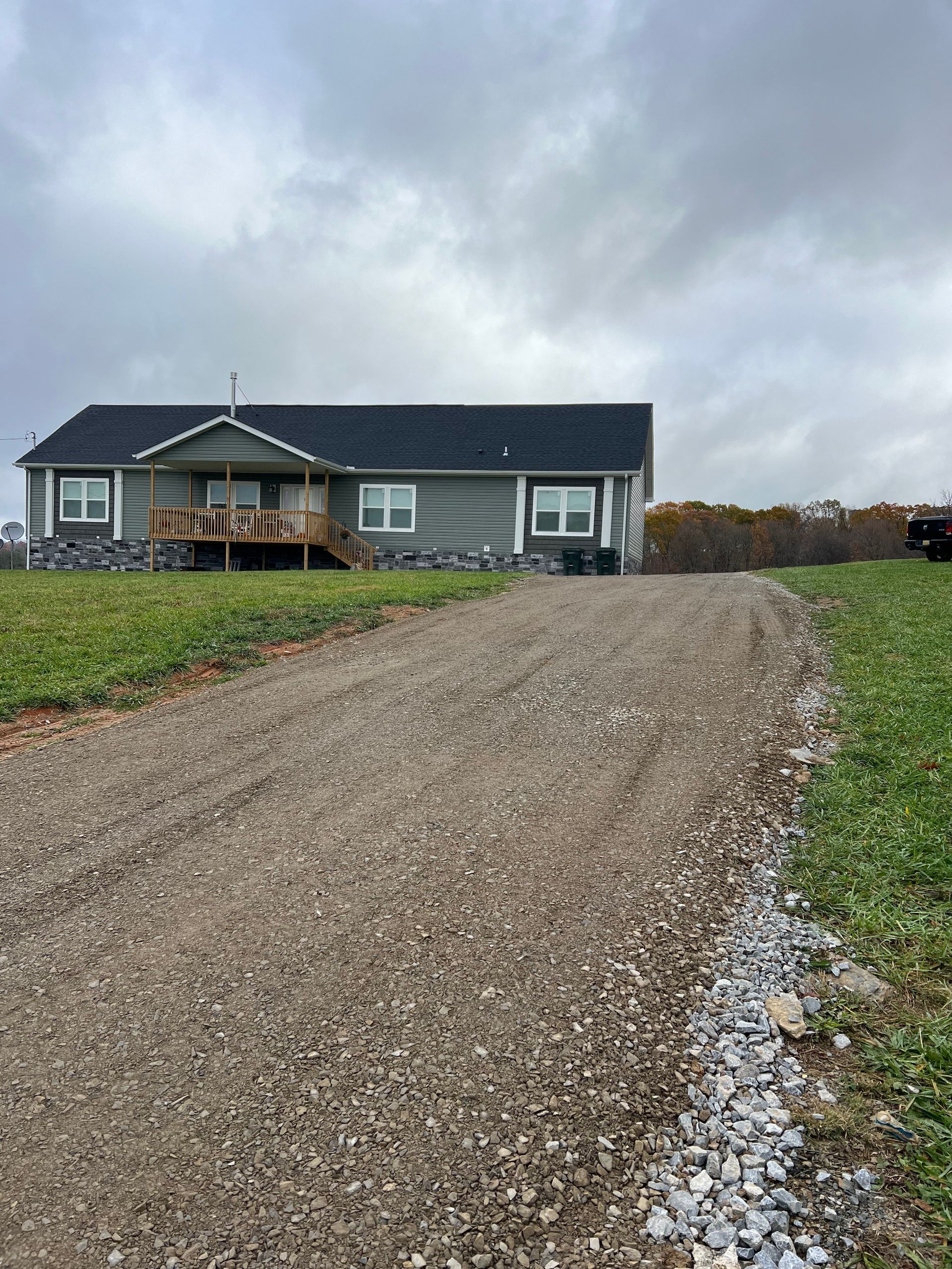 A gravel driveway leads to a gray house with a black roof under an overcast sky.