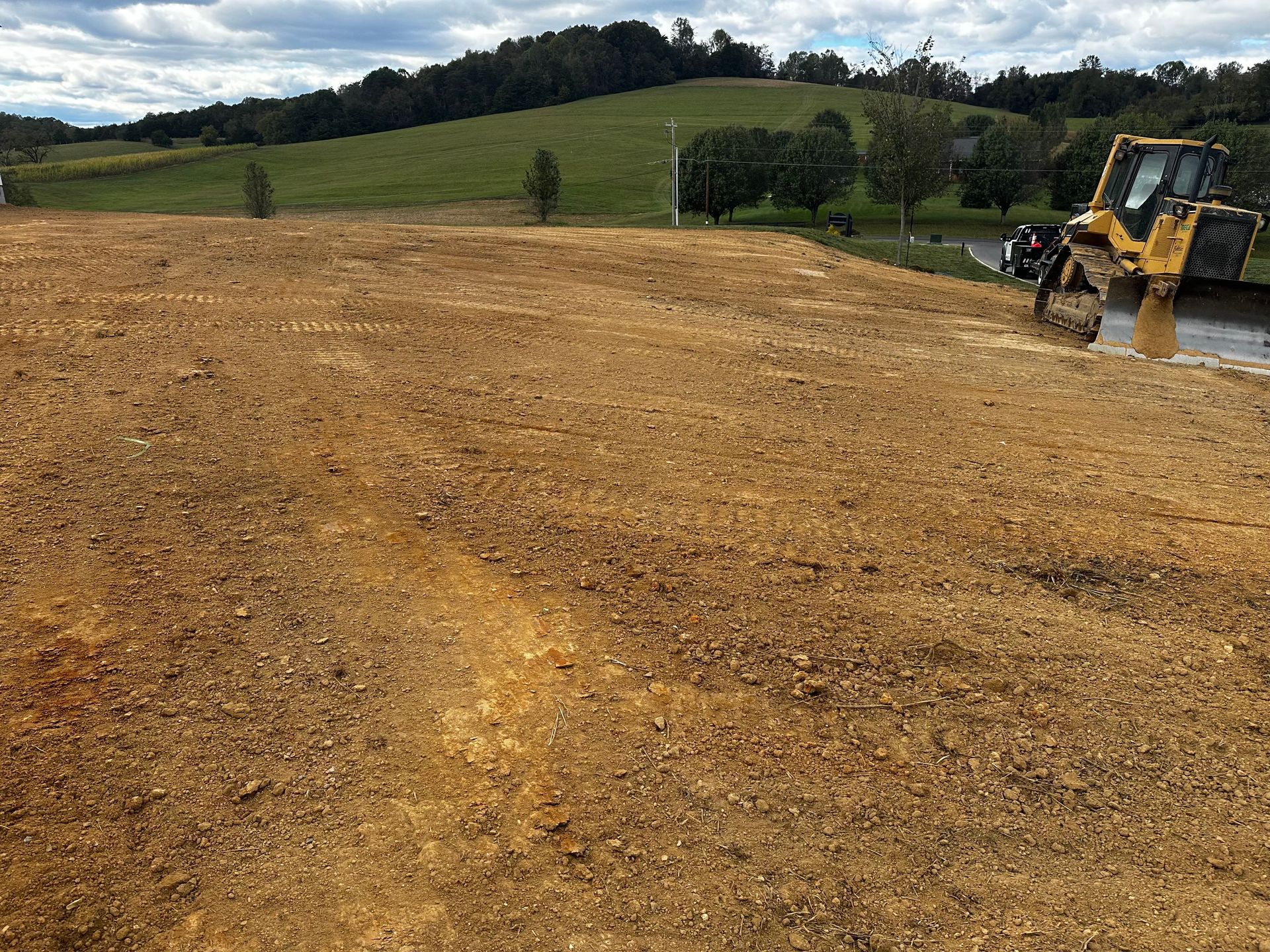 Yellow bulldozer on dirt site, preparing land. Green hill in background. Overcast sky.