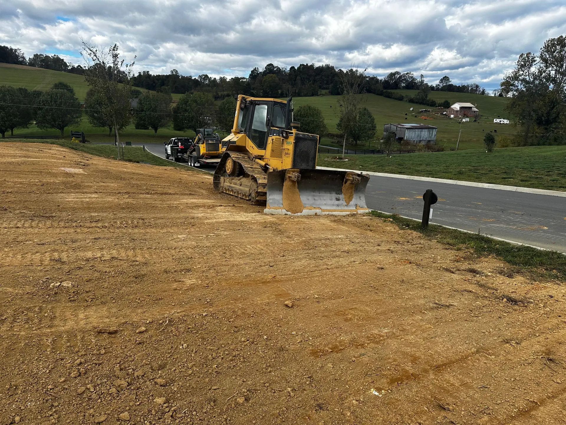 Yellow bulldozer on a dirt lot next to a road, leveling the ground. Green hills in the background.
