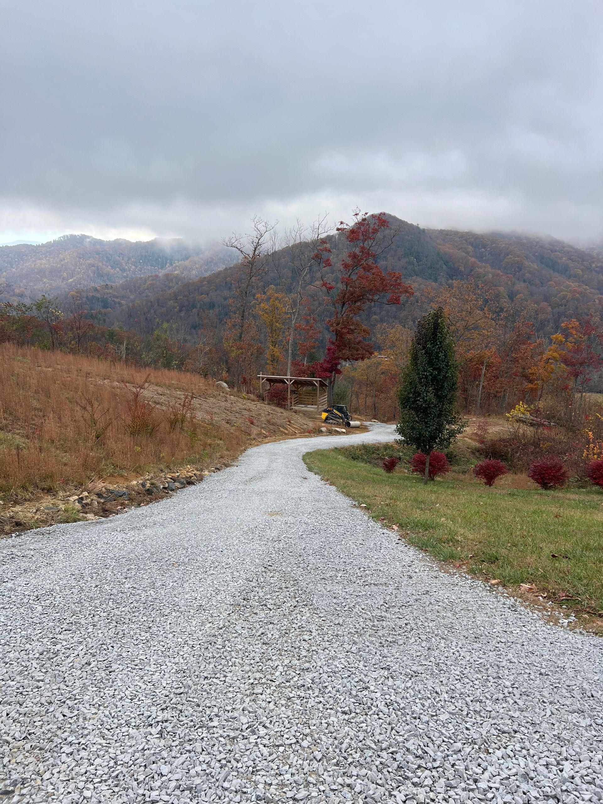 Gravel driveway leading to a mountain under cloudy sky. Autumn colors in trees, grass.