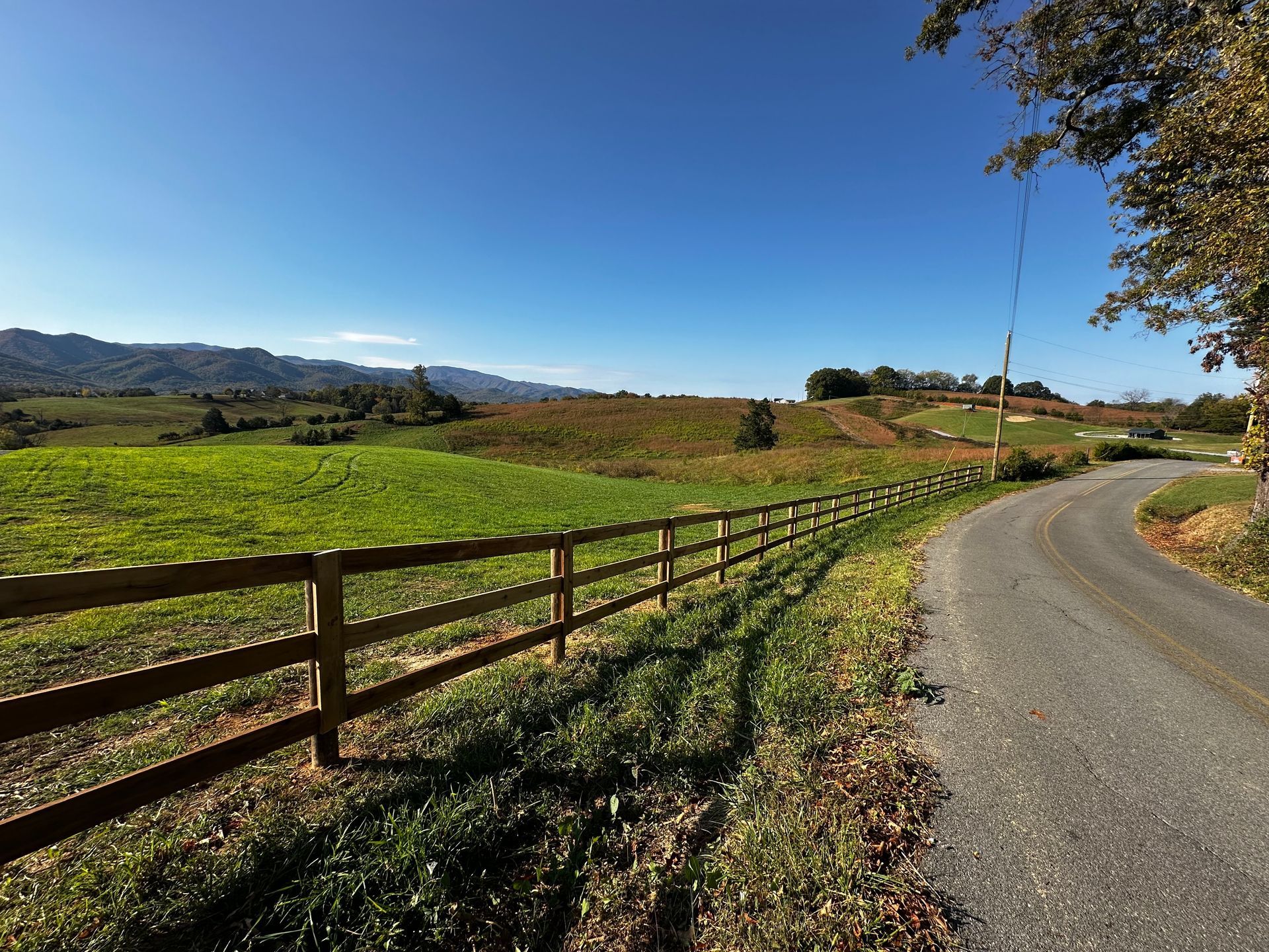 A rural landscape with a wooden fence, green field, and winding road under a blue sky.