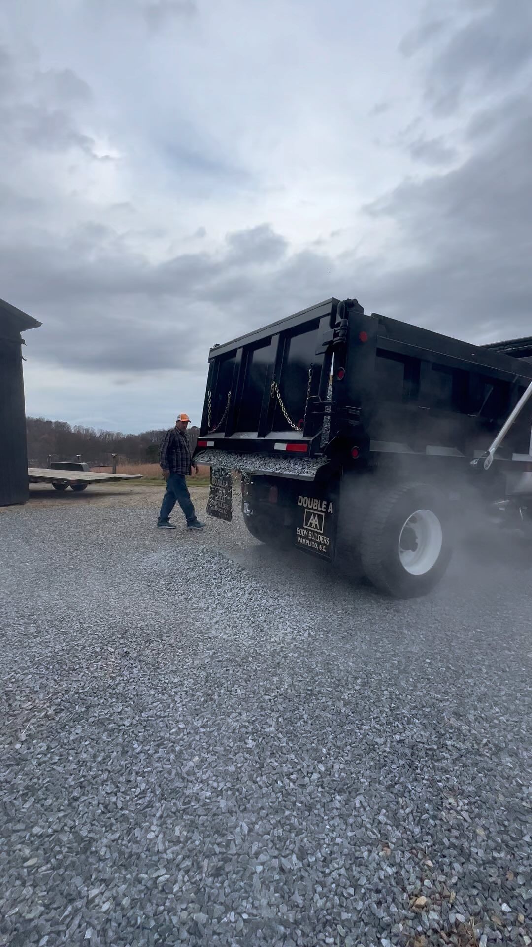 Man operating a black dump truck on a gravel surface on a cloudy day.