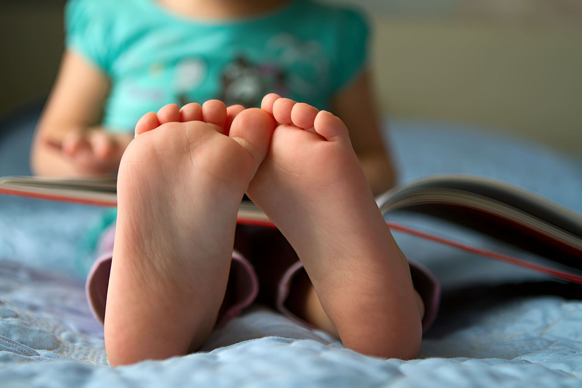 A Little Girl Is Sitting On A Bed Reading A Book — Hastings Podiatry Centre In Port Macquarie, NSW