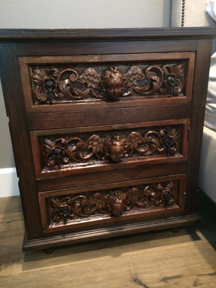 A carved wooden nightstand with three drawers is sitting on a wooden floor next to a bed.