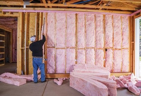Man installing pink insulation in a wooden-framed wall, inside a house under construction.