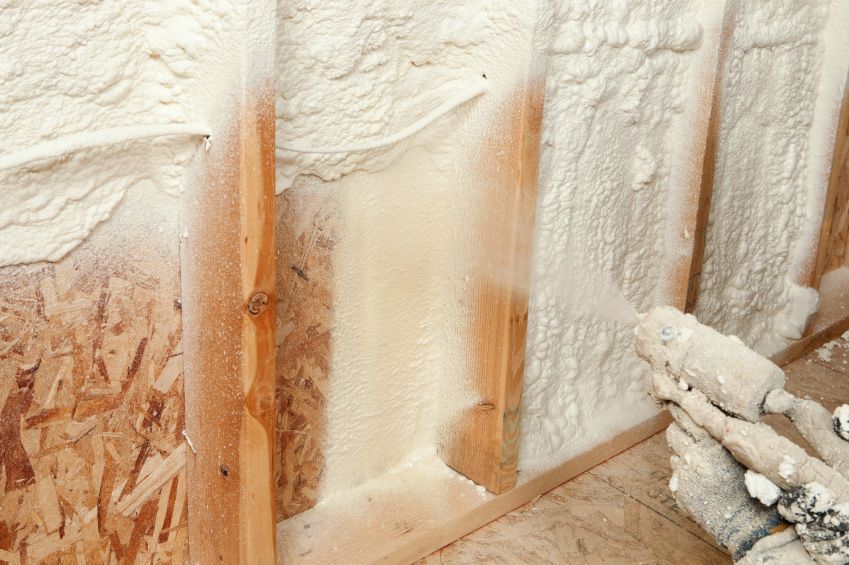 Foam insulation being sprayed into a wall cavity between wooden studs, creating a white barrier.