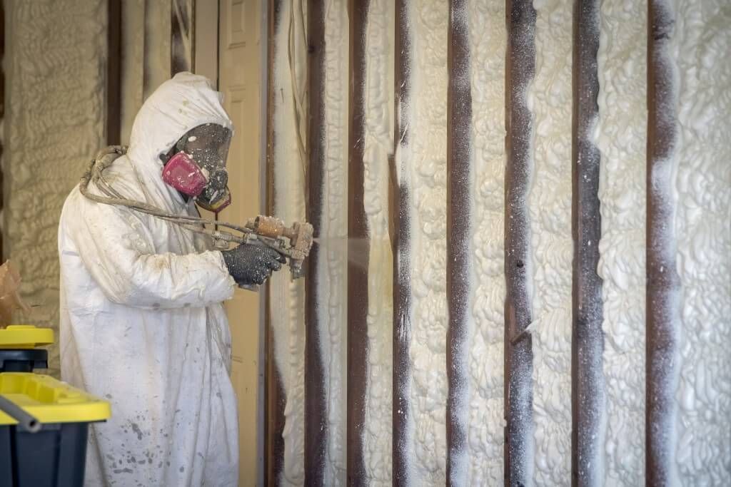 Person in protective suit sprays insulation onto a wooden wall.