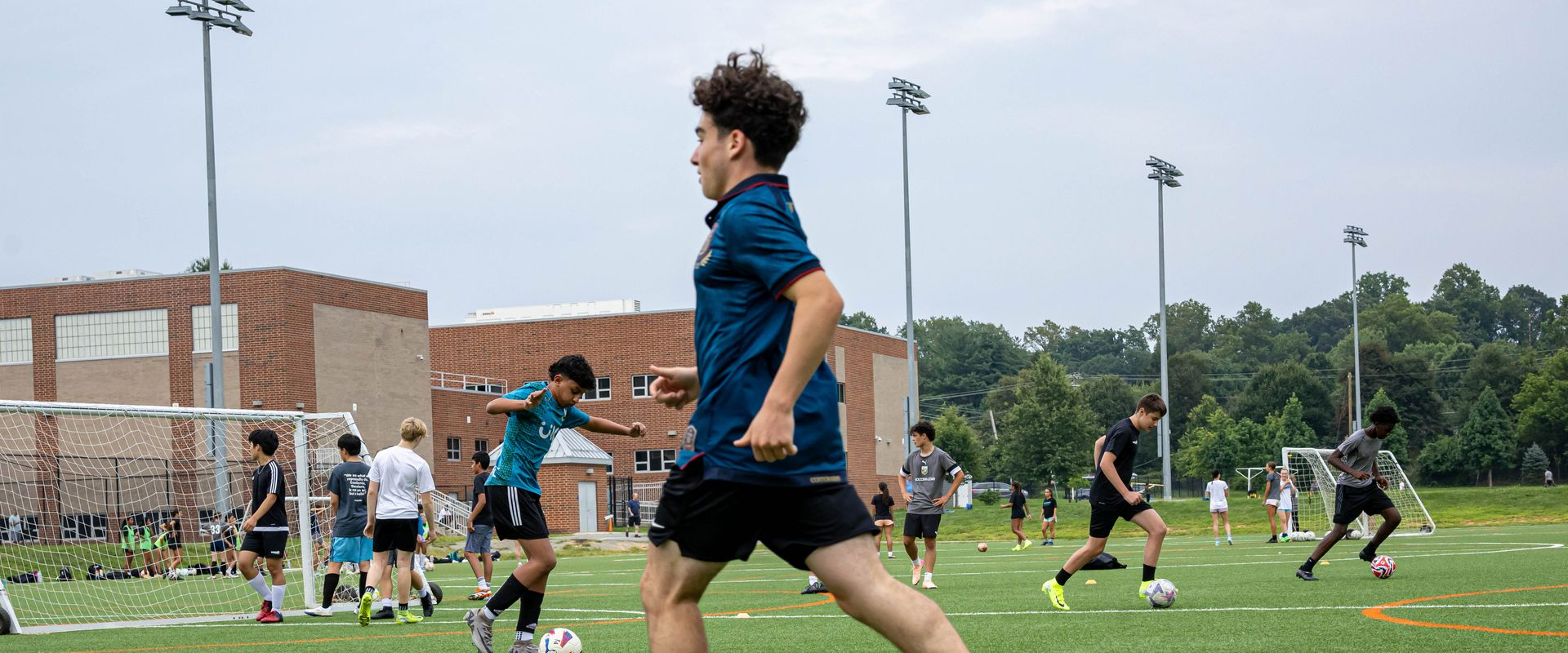 MSI Soccer teens dribble through ball-control drills on a turf field beside a school, with cones and goals set up around the session