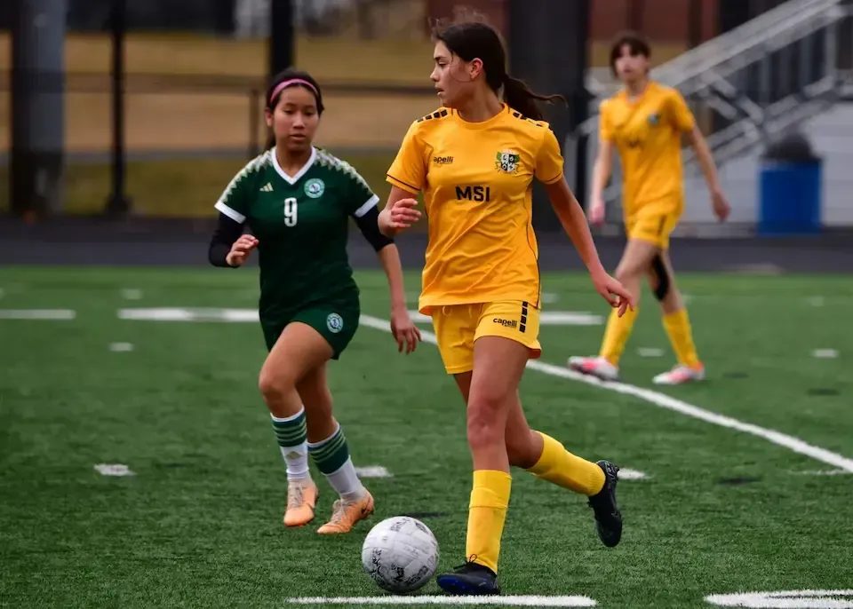 MSI soccer player in yellow uniform controls the ball while being defended by an opponent in green during a competitive match.