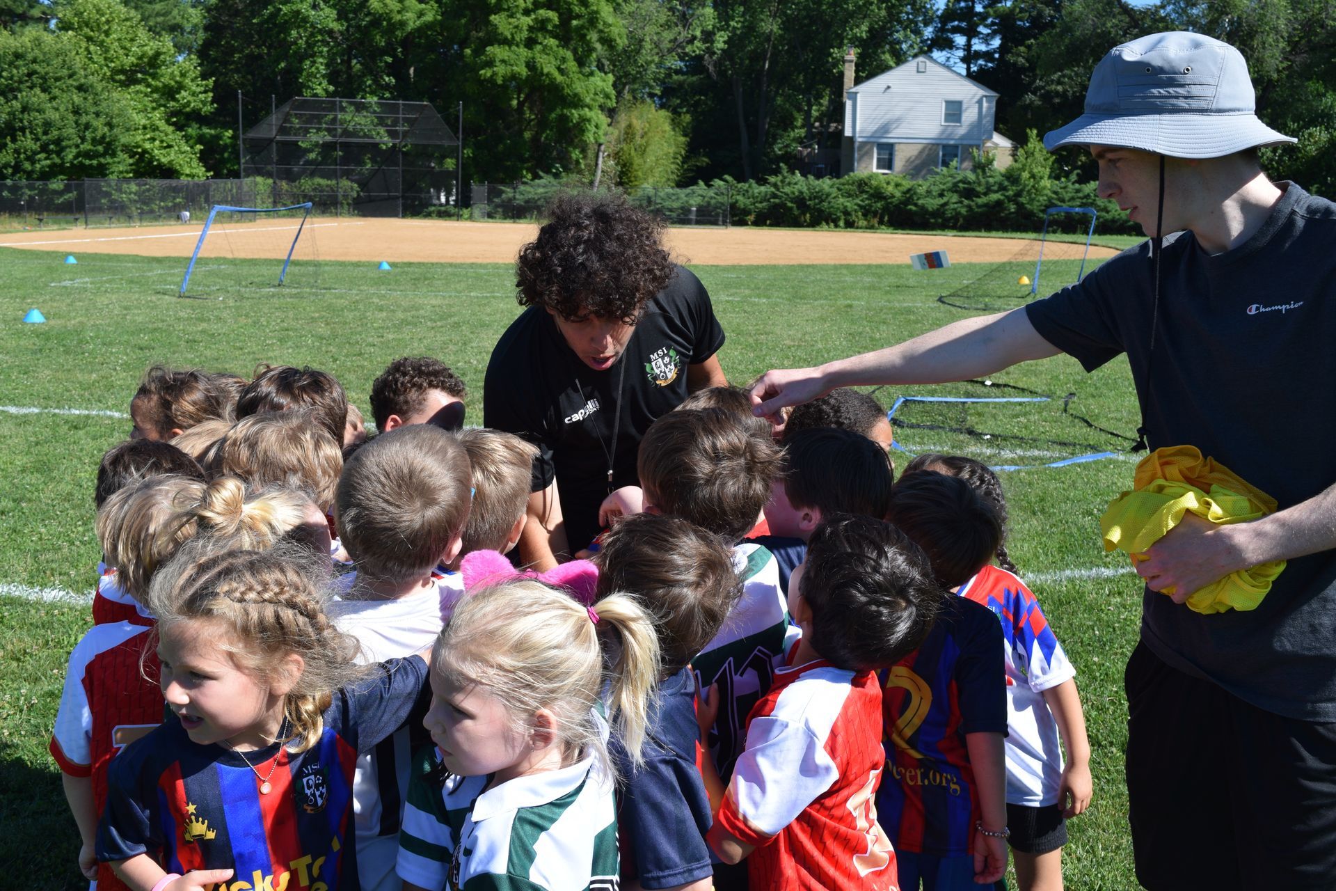Children in MSI soccer jerseys gather around coach on sunny field, listening to instructions and preparing for practice.