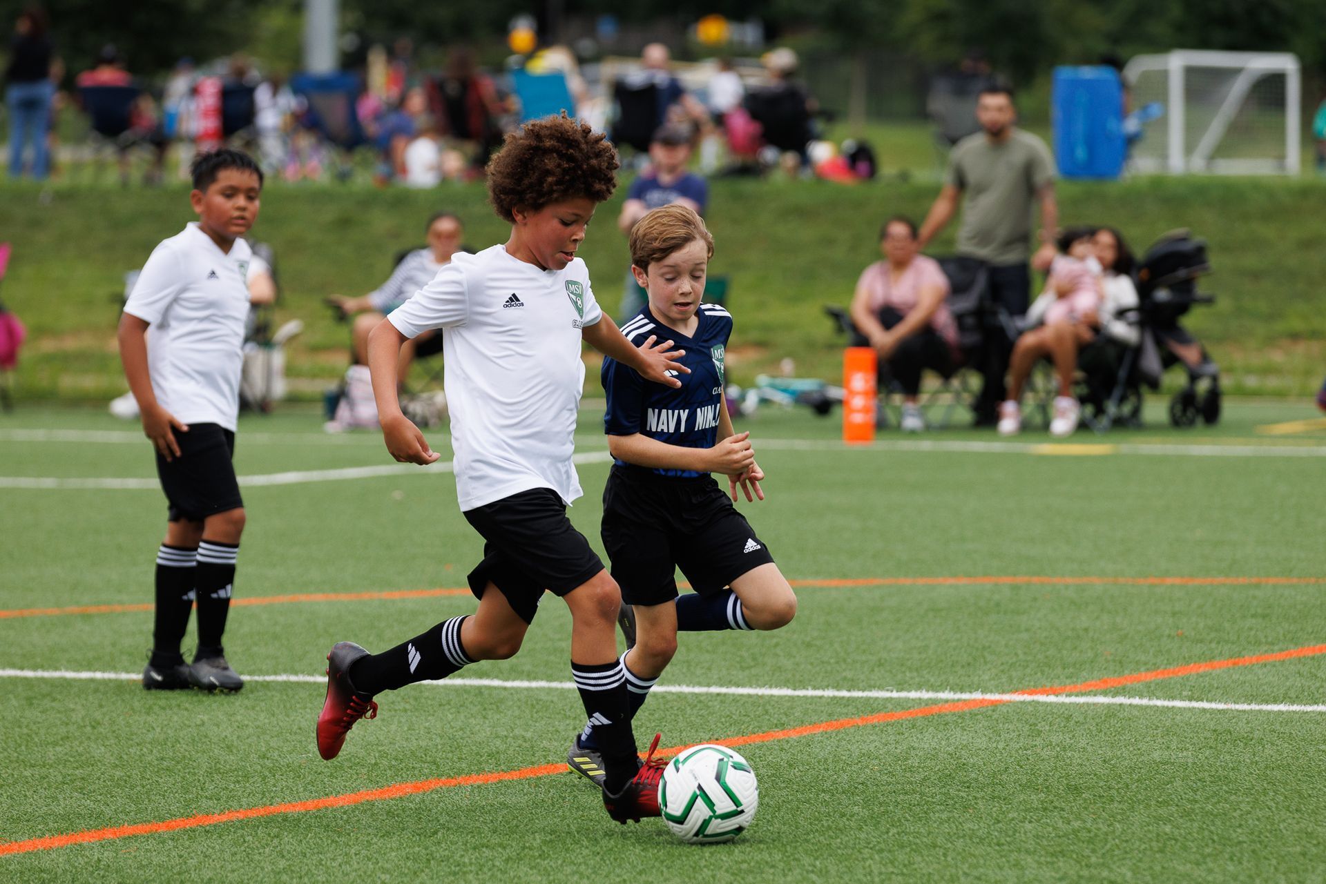 Two MSI youth players battle for the ball during a Classic League game on a turf field, with families watching from the sideline in Montgomery County, MD.