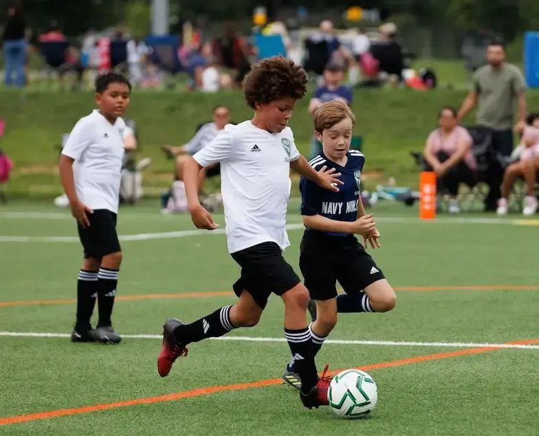 Two MSI youth players battle for the ball during a Classic League game on a turf field, with families watching from the sideline in Montgomery County, MD.