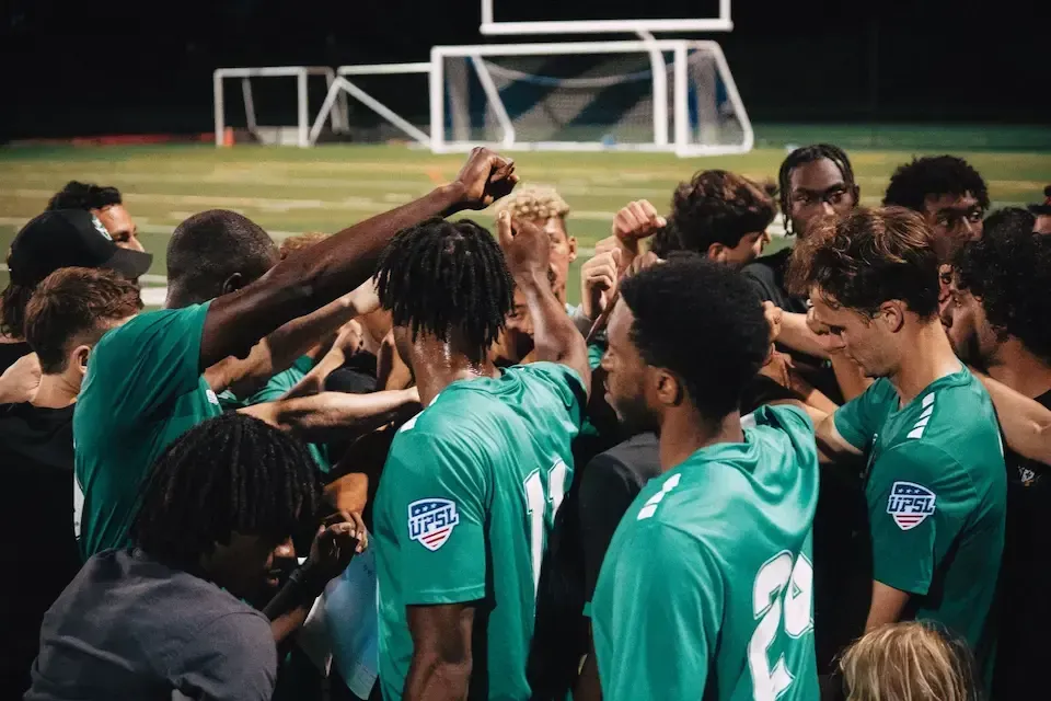 Players from the MSI soccer team unite in a powerful pre-game huddle, wearing green uniforms with UPSL league patches.