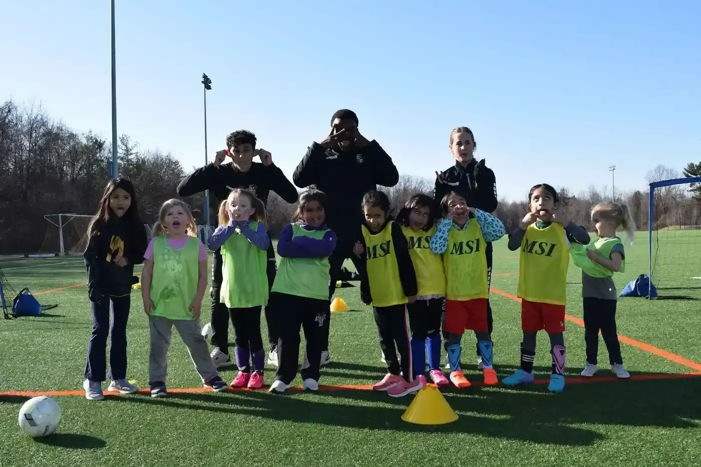 MSI soccer kids in green pinnies pose with coaches on turf field, making silly faces and having fun during practice.