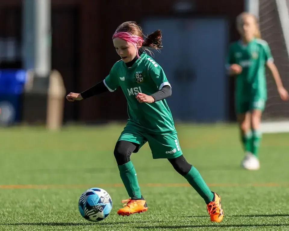 Girl in MSI green soccer uniform running and kicking a soccer ball during a soccer match