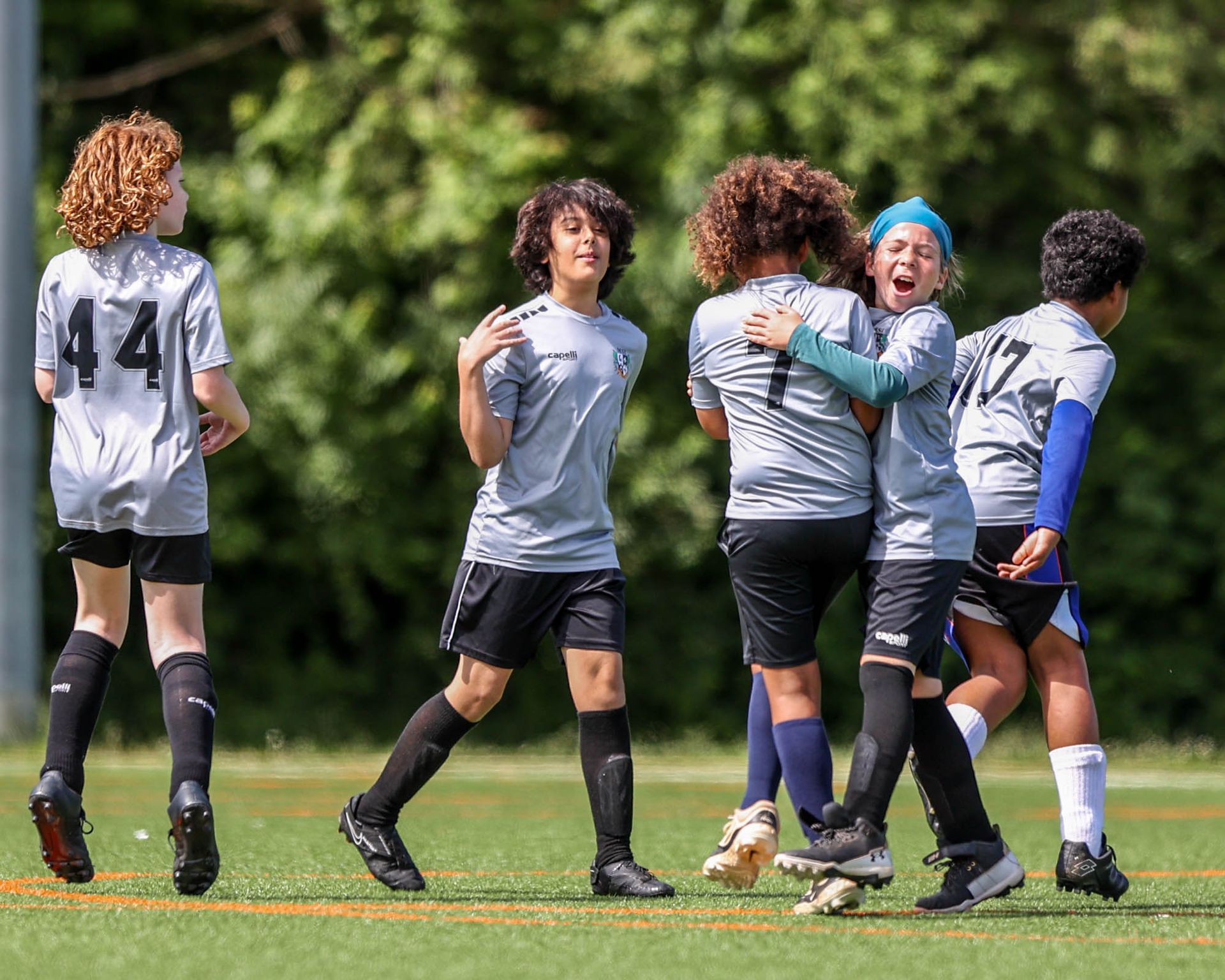 MSI soccer players in gray uniforms hug and smile in celebration on the field after scoring a goal in a youth soccer match.