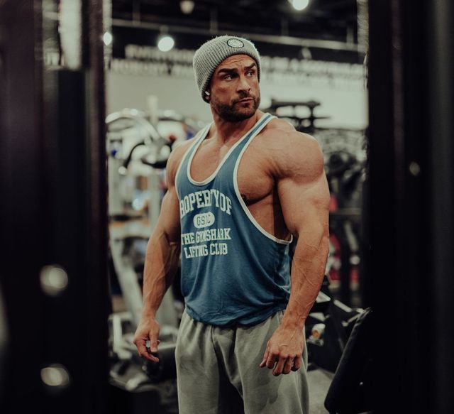 A muscular man wearing a tank top and a beanie is standing in a gym.