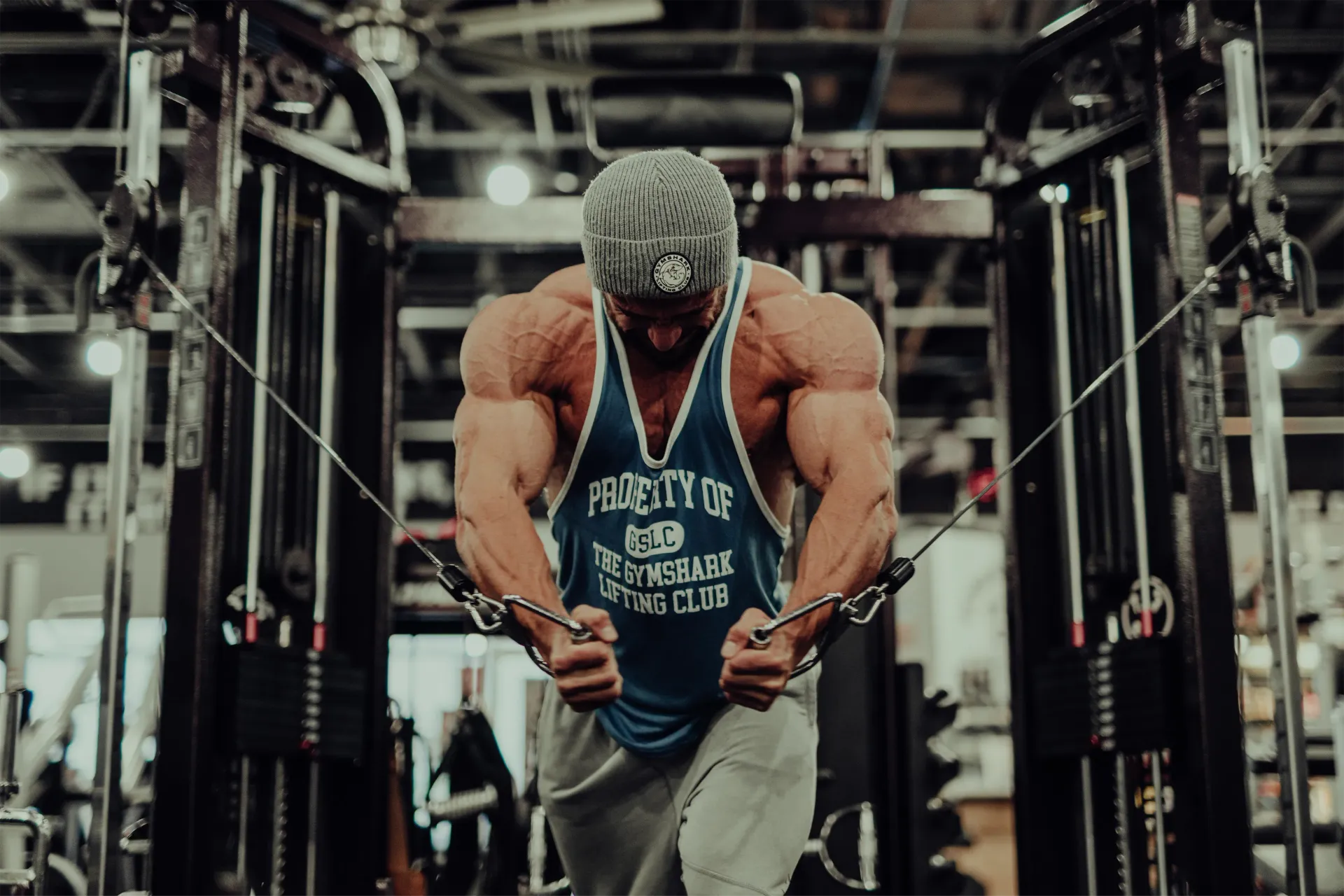 A muscular man wearing a tank top and a beanie is standing in a gym.
