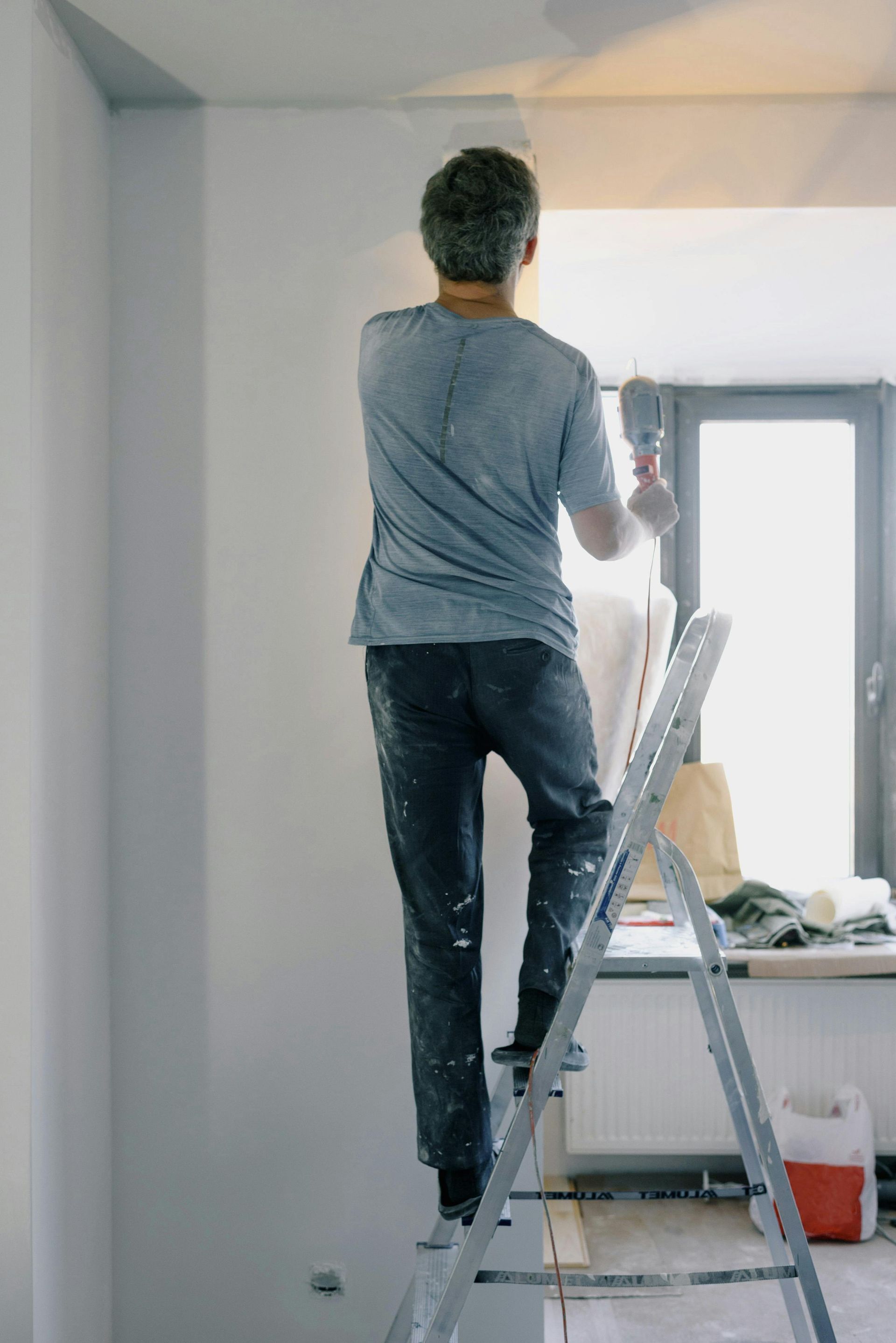 Man on ladder, painting a white wall near a window.