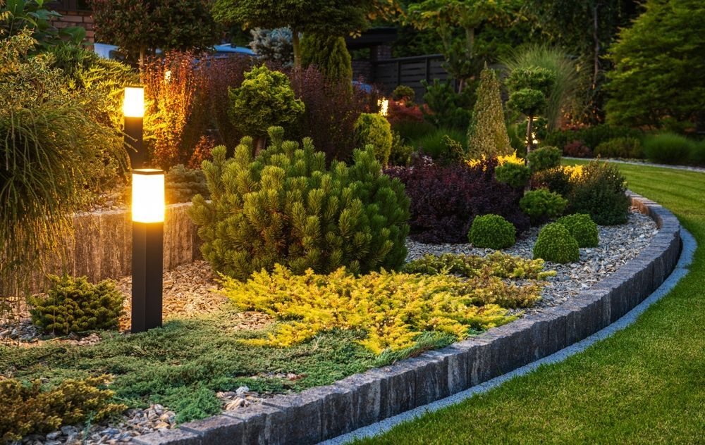 Illuminated garden bed with various green and yellow plants, lined with stone edging, and lawn.