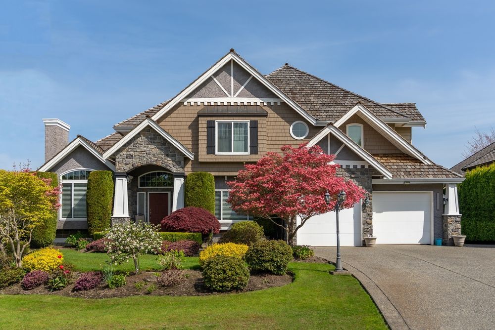 Two-story house with brown and gray exterior, lush green lawn, and vibrant landscaping.