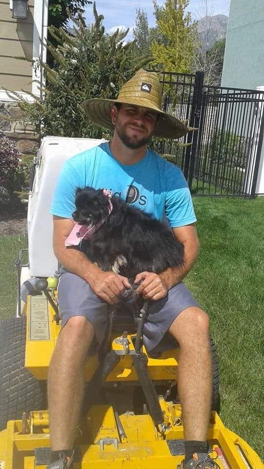 Man in straw hat and blue shirt sitting on lawnmower, holding a black dog on a sunny lawn.