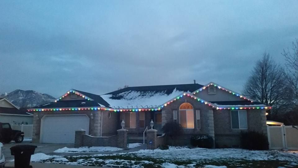 House with Christmas lights on the roof, snow on the ground and a mountain in the background.