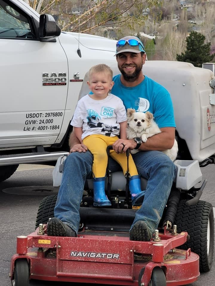 Man, child, and small dog sit on a red lawnmower in front of a truck.