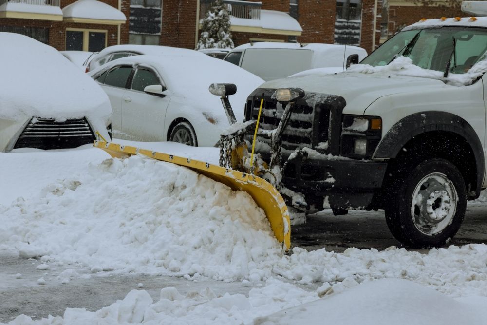 Snowplow clearing snow from a street, with parked cars covered in snow in the background.
