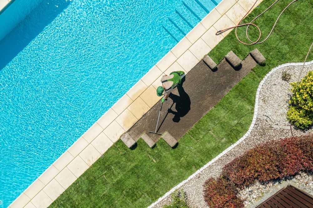 Person rolling out sod next to a pool. Rolls of sod on a strip of grass. Lush greenery and blue water.