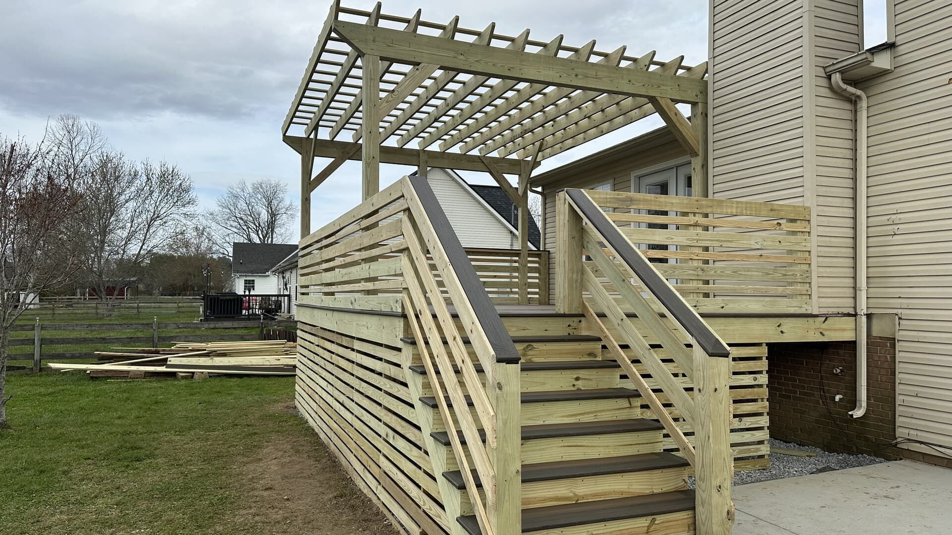 Wooden deck with pergola and stairs next to a house; cloudy sky.