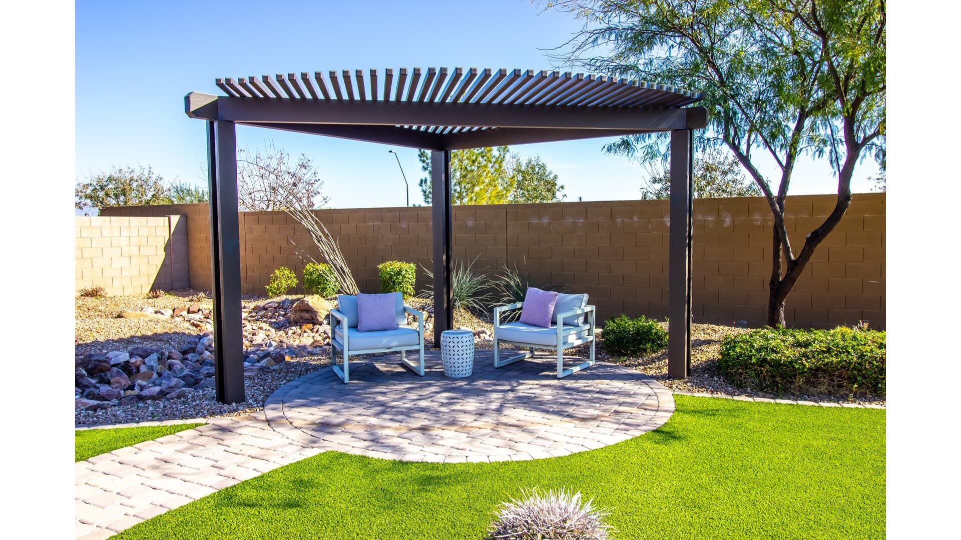 Pergola with seating area on a stone patio in a yard with green grass and a neutral-colored wall.