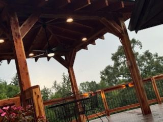 Wooden gazebo with lighting and ceiling fan, overlooking a deck with trees and a cloudy sky.
