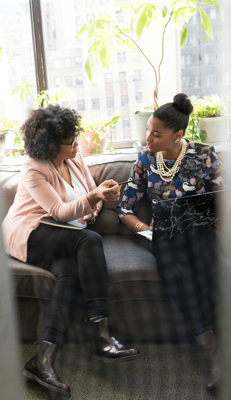 Two women sitting on a couch, talking. One wears a pink blazer, the other a patterned top. Sunlight streams in.