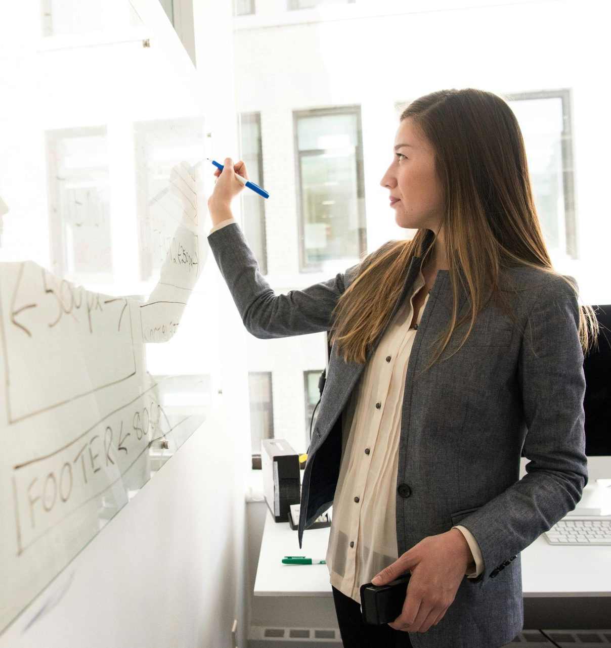 Woman in blazer writing on whiteboard, office setting.