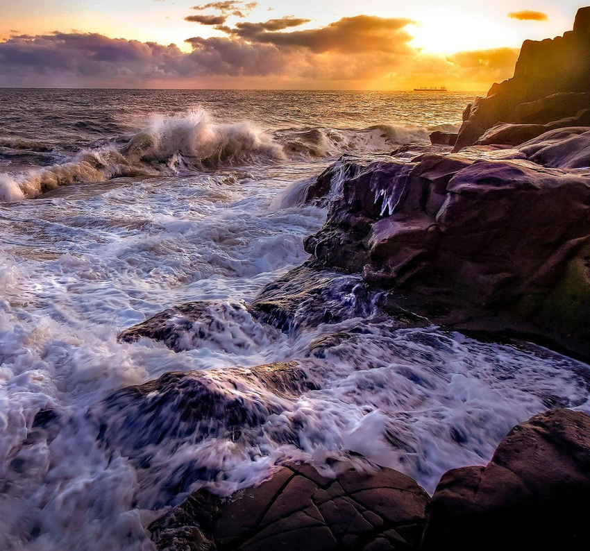 Waves crash on rocky shore at sunset, with golden light reflecting on water and clouds.