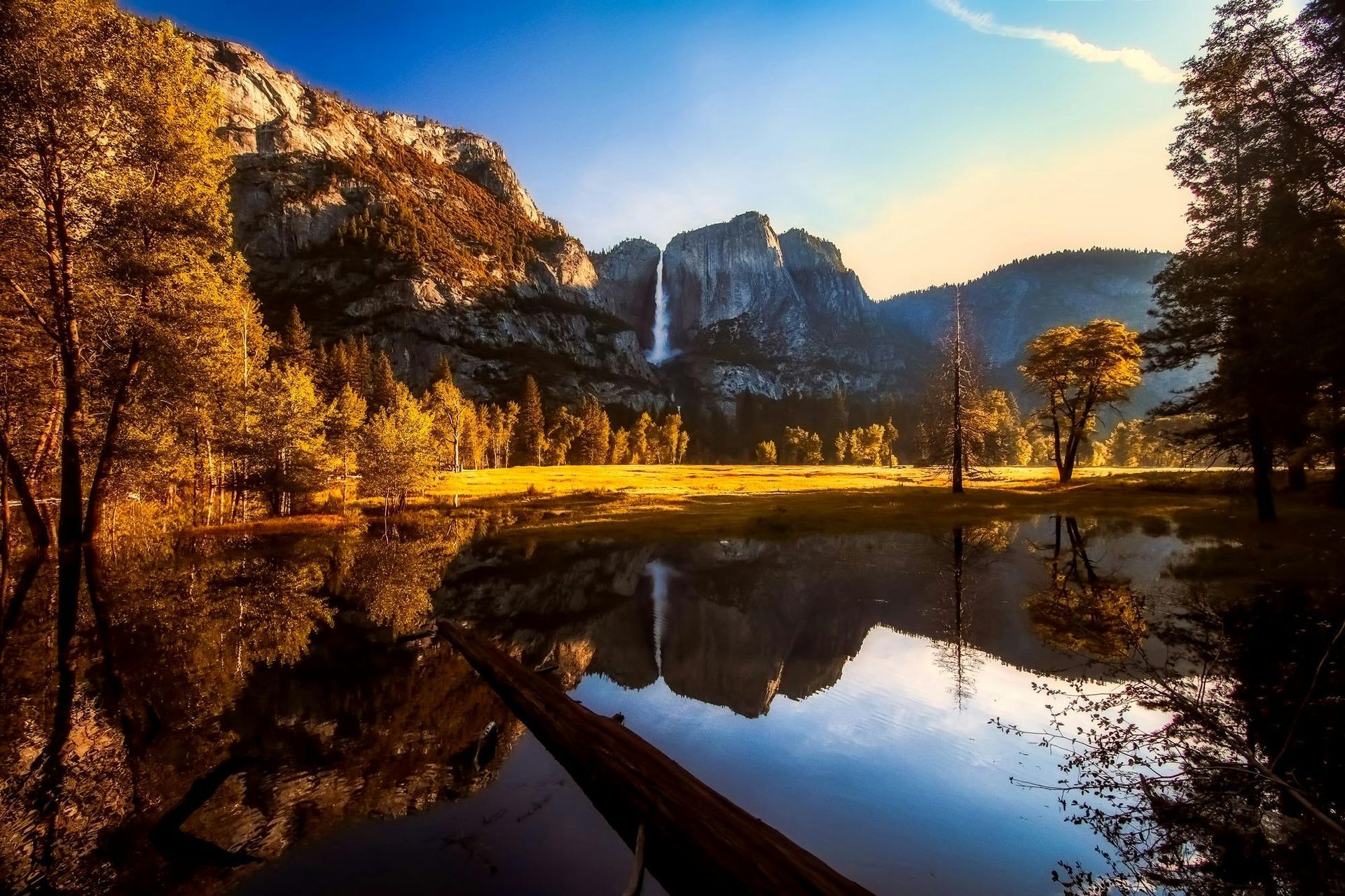 Mountains and waterfall reflect in a calm lake; autumn trees in the foreground; blue sky.
