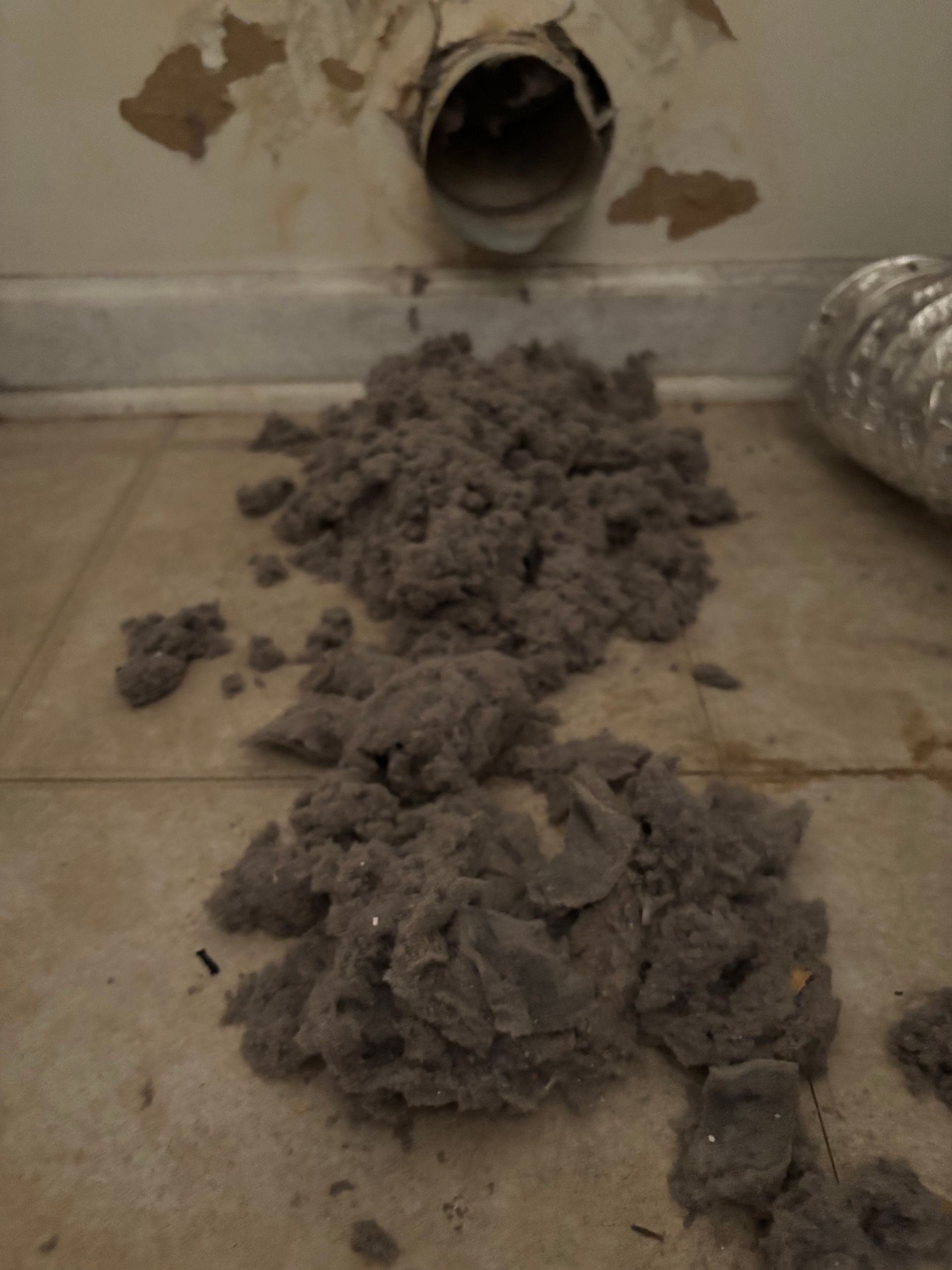 A pile of dryer lint coming out of a dryer vent pipe on the floor.