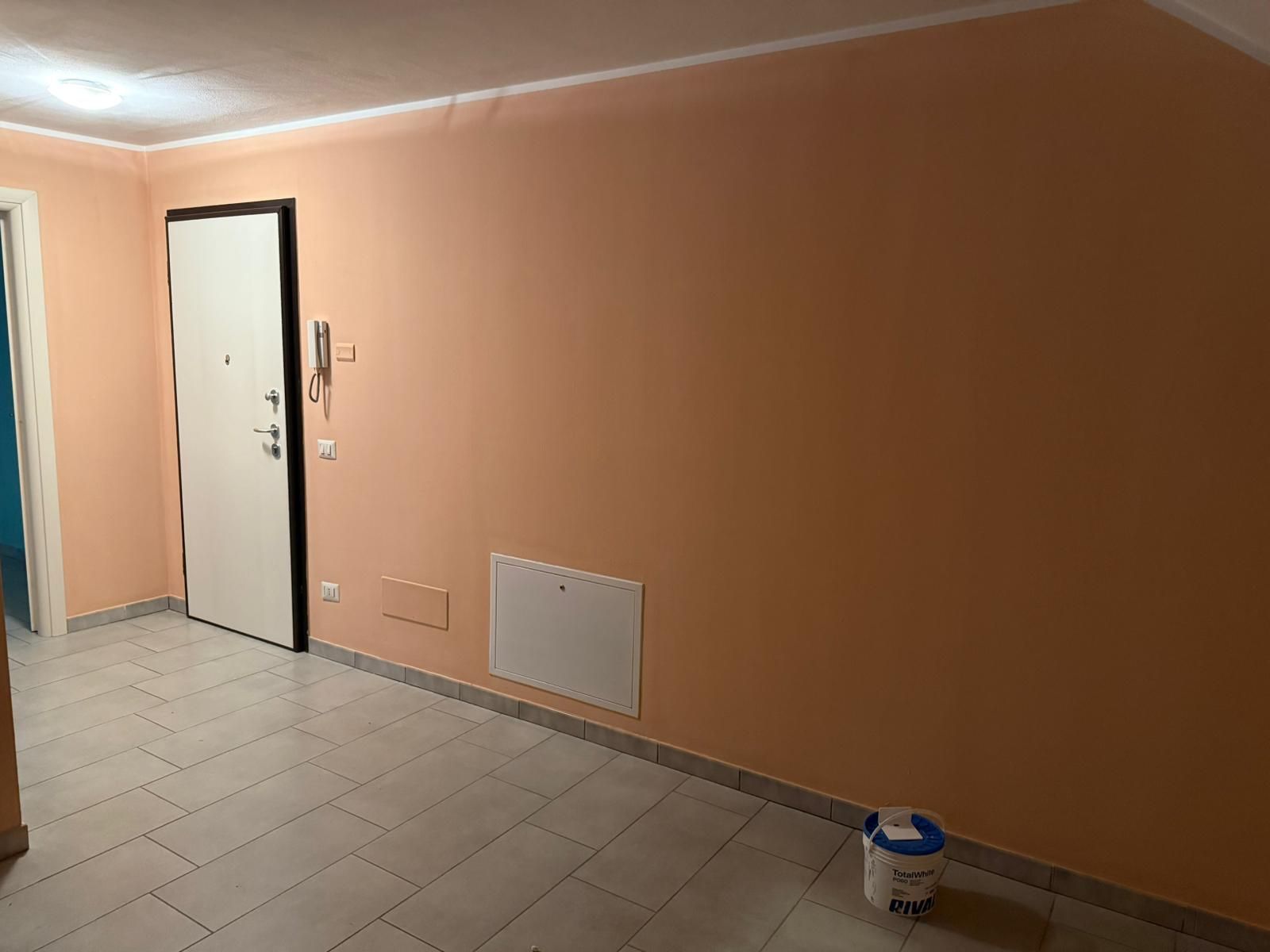 Corridoio vuoto con pareti color pesca, porta bianca, pavimento piastrellato e un secchio di vernice vicino al muro.