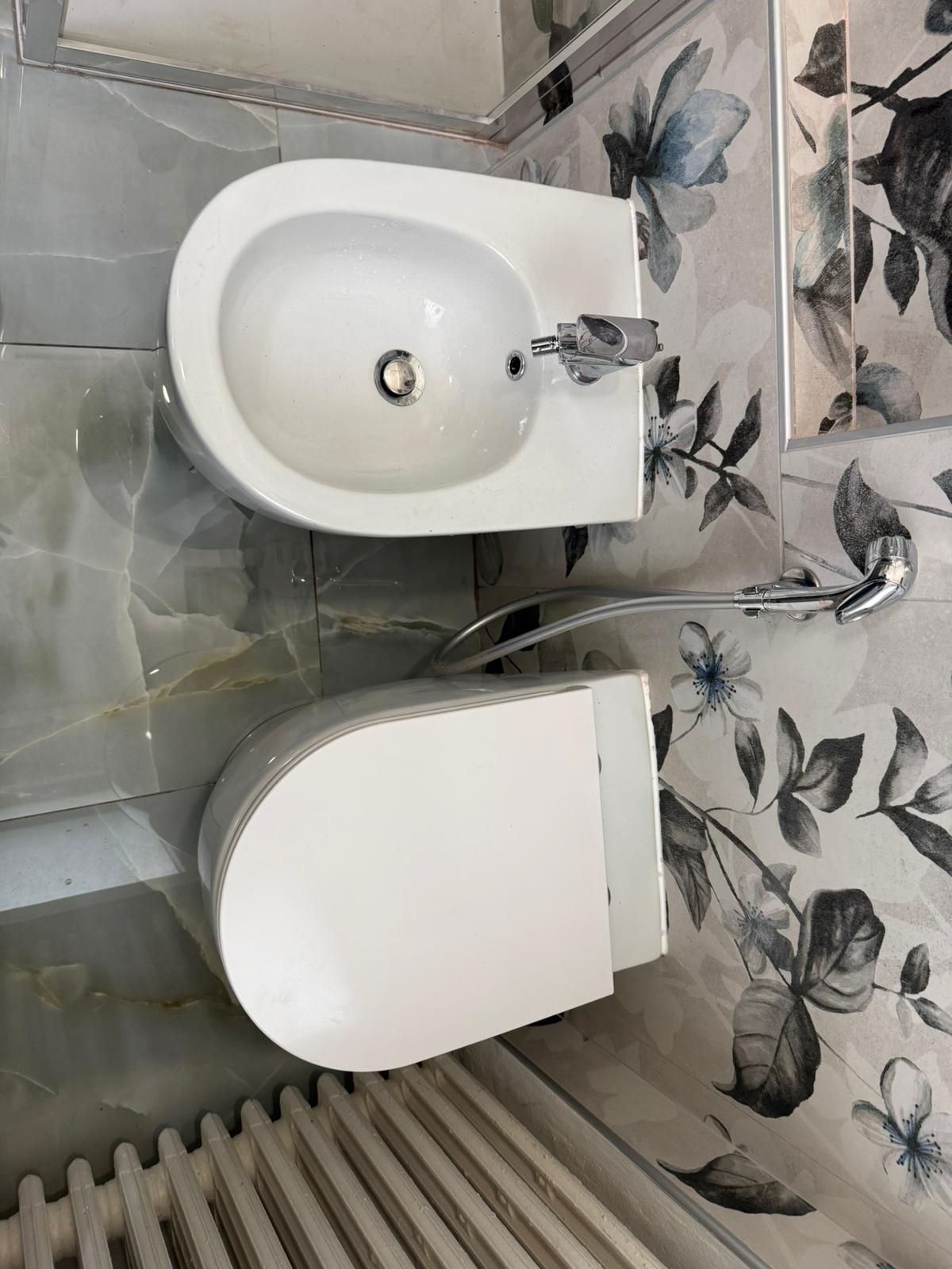 Piccolo bagno con WC bianco, lavandino, termosifone, piastrelle grigie e carta da parati con motivo a farfalle.